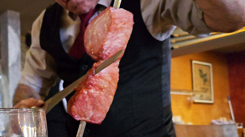 Bem Brasil In Deansgate Manchester - A waiter in a vest serves grilled meat on a skewer. He's slicing it with a knife. A glass and wooden interior are visible in the background.