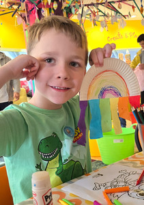 We Invented The Weekend Festival - Smiling boy in green dinosaur shirt holds a paper rainbow craft at a colorful art table in a bright playroom.
