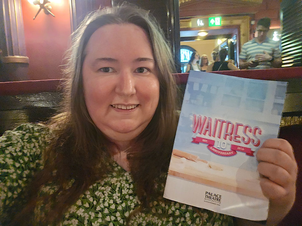Smiling woman in a theater holds a Waitress 10th anniversary program; Palace Theatre Manchester visible behind her.
