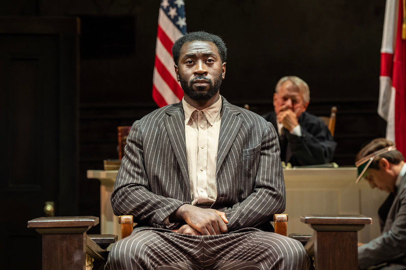 To Kill a Mockingbird - Man in striped suit sits solemnly in courtroom, flanked by American flags. Judge and note-taker present in background, creating a tense mood.