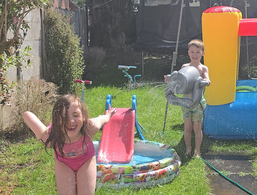 Two kids play in a sunny backyard splash pool, one spraying water from a gray toy while a girl in pink laughs near a red slide.