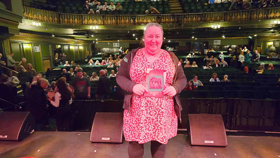Woman in red dress holds "Choir of Man" program on stage. Theater audience is visible in the ornate venue. Warm lighting creates a lively mood.