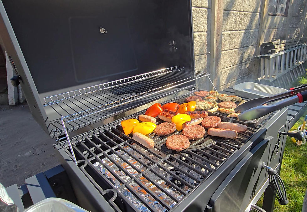 Grill with sausages, burgers, and colorful bell peppers in a sunny backyard. Tongs turning the food. Stone wall and grass in background.