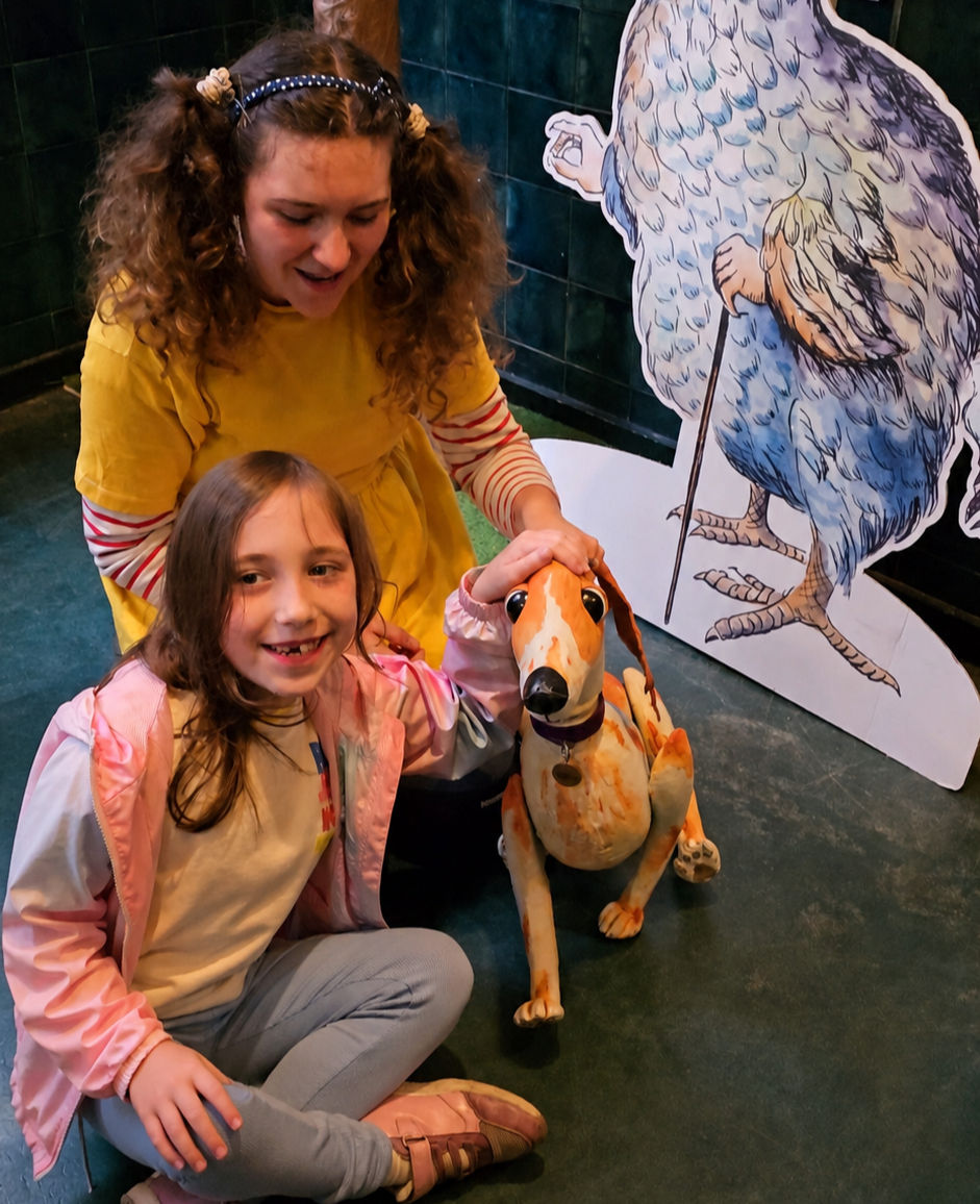 Girl and woman in colorful clothing pet a dog sculpture indoors. A large cartoon bird cutout stands in the background. Both are smiling.