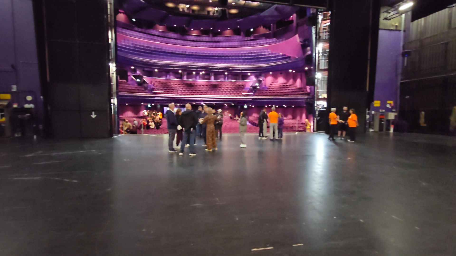 Lowry Backstage Tour - People gather on a theater stage floor before empty purple seats and balconies, with staff in orange shirts in a dim auditorium.