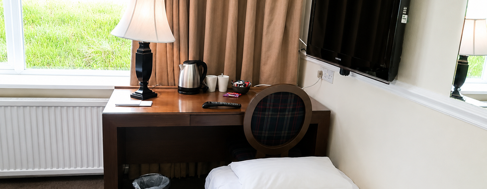 Family room at Hollin House Hotel showing a single camp bed beside a wooden desk with tea and coffee facilities, wall-mounted TV, lamp and large window with curtains.