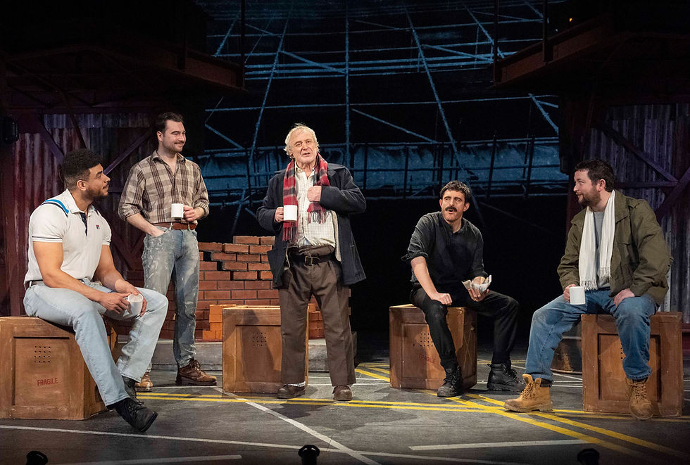 Five men in casual attire sit on crates, each holding a mug, on a stage with a brick wall backdrop. The scene is lively and conversational.