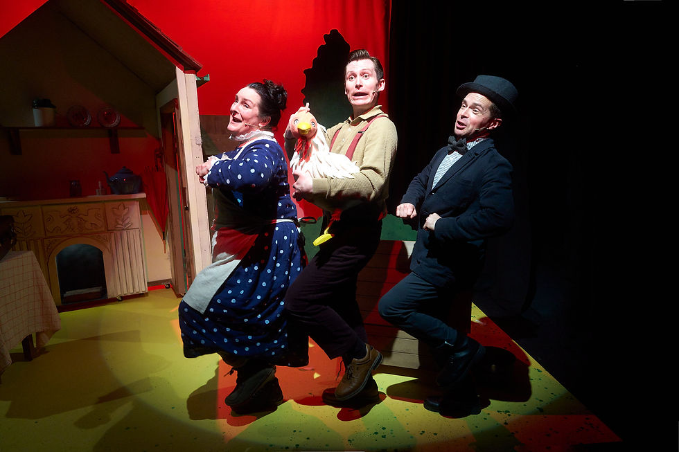 A Squash and a Squeeze uk tour - Three people in playful poses, one holding a chicken toy, inside a colorful set resembling a house. Bright lighting and joyful expressions.