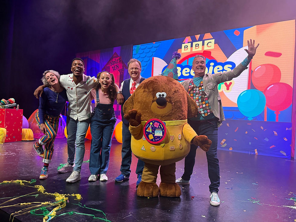 CBeebies House Party Live! -Group of five people and a costumed character posing joyfully on stage with colorful balloons and "BBC CBeebies" backdrop.
