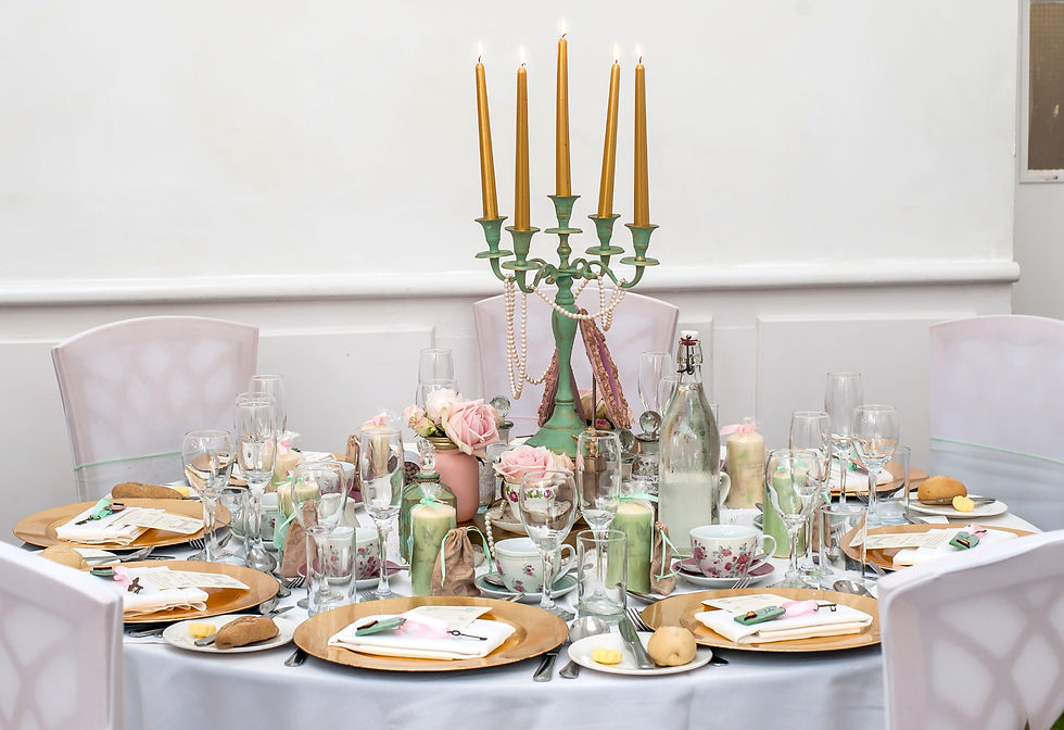Elegant dining table set with lit gold candles, pink roses, glassware, and floral cups in a bright white room