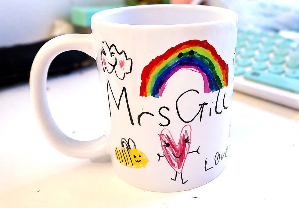 White mug with a hand-drawn rainbow, heart, bee, and cloud labeled Mrs Gill and Love on a desk.