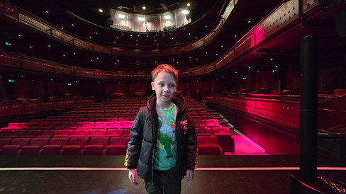 Lowry Backstage tour - Young boy stands on a theatre stage before red seats and balcony levels, lit by pink and warm lights, looking curious and calm.
