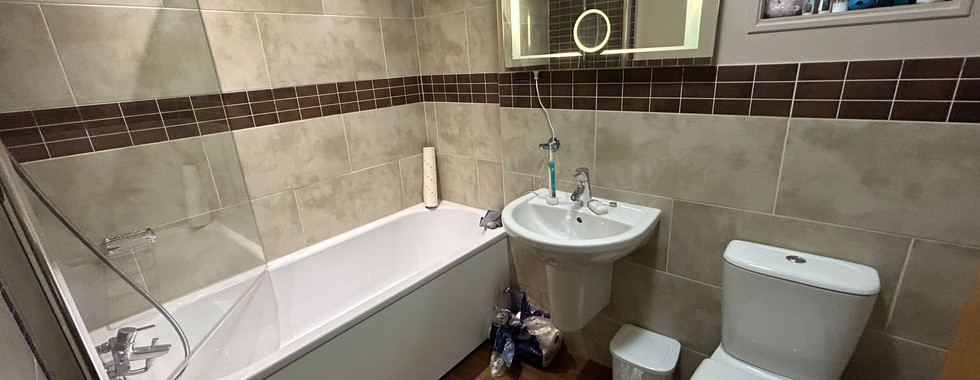 Modern family bathroom in the Diamond Cottage featuring a bath with overhead shower, illuminated mirror, toilet, and sink.