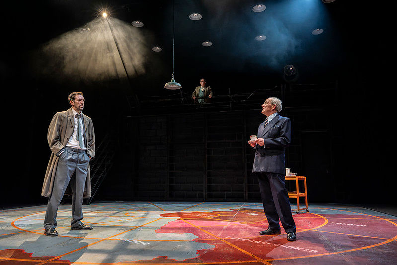 Ralf Little as Alec Leamas faces Nicholas Murchie as Control on the minimalist set of The Spy Who Came in from the Cold, with Tony Turner as George Smiley watching from an elevated platform.