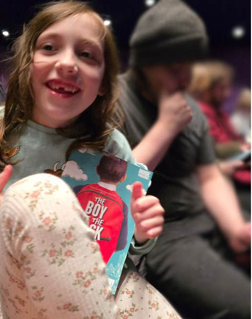 Girl smiling and holding program titled "The Boy in the Dress." She sits in a theater, wearing floral pants; background is blurred.