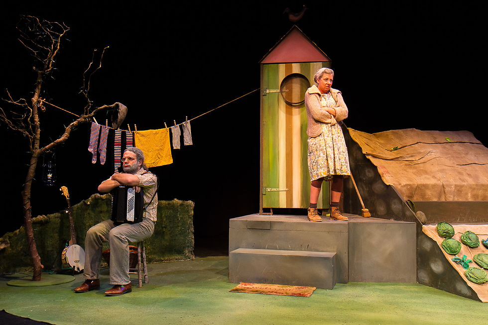 The Enormous Turnip at Waterside Arts - A man with an accordion sits near clothes on a line; a woman stands by a striped shed, arms crossed, in a garden setting with earthy tones.