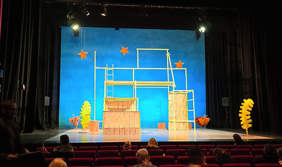 Stage with blue backdrop, yellow scaffolding, orange stars, and abstract props. Audience seated in red chairs, theater setting.