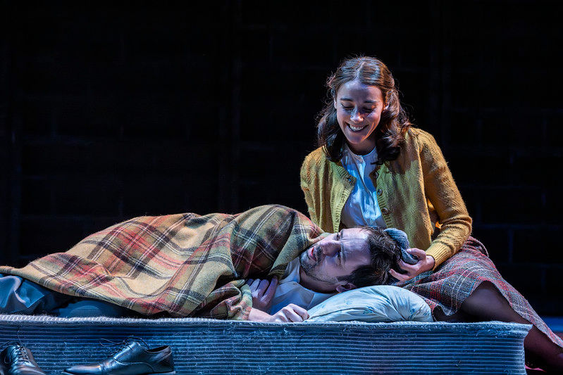 Ralf Little as Alec Leamas rests with his head on Liz Gold's lap as Gráinne Dromgoole smiles beside him during The Spy Who Came in from the Cold.