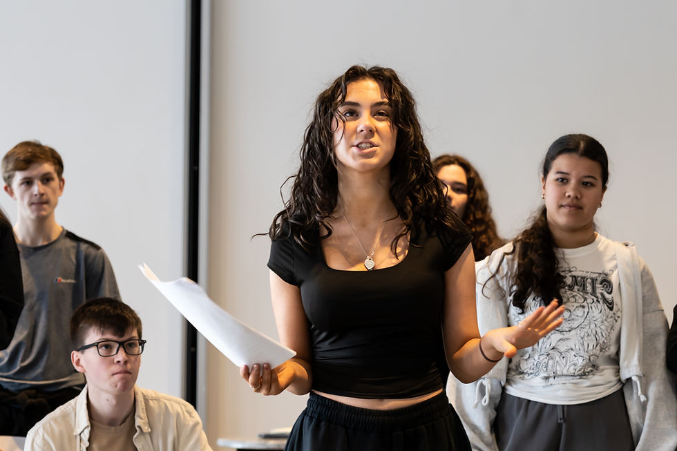 Young woman in black top speaks confidently, holding papers in front of a small group in a bright room. Others listen attentively.