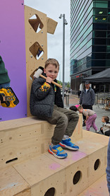 We Invented The Weekend Festival - Boy in black jacket sits on a wooden play structure holding a mallet, with other children in an urban plaza.