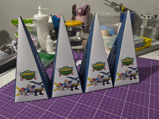 Quatro caixas piramidais de papel personalizadas com personagens do Sonic (Tails, Amy, Big e Sonic). As laterais azuis exibem o texto "AUGUSTO 6 ANOS".