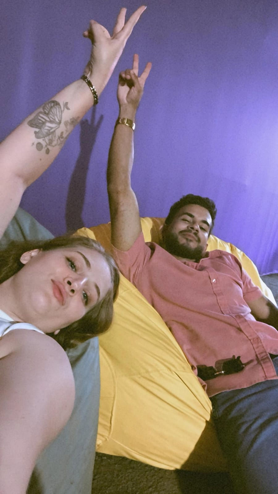  Uma selfie tirada de cima mostra uma mulher loira e um homem de barba deitados em um ambiente com uma parede roxa vibrante. A mulher, em primeiro plano, tem um braço levantado fazendo um sinal de "paz" e uma tatuagem de borboleta no antebraço. O homem, atrás dela, está em um pufe amarelo e também levanta um braço fazendo um sinal de "chifres". Ambos parecem relaxados e olham para a câmera.