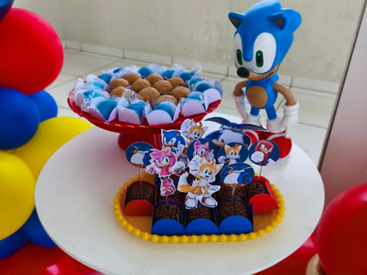 Visão de uma mesa redonda branca com docinhos variados. Há uma bandeja amarela com brigadeiros decorados por toppers de Amy, Tails e Knuckles, e uma boleira vermelha ao fundo. Ao lado, destaca-se um boneco grande de plástico do Sonic.