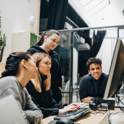 image of one woman working on a computer with several other people standing around her watching what she's doing - an image to represent training