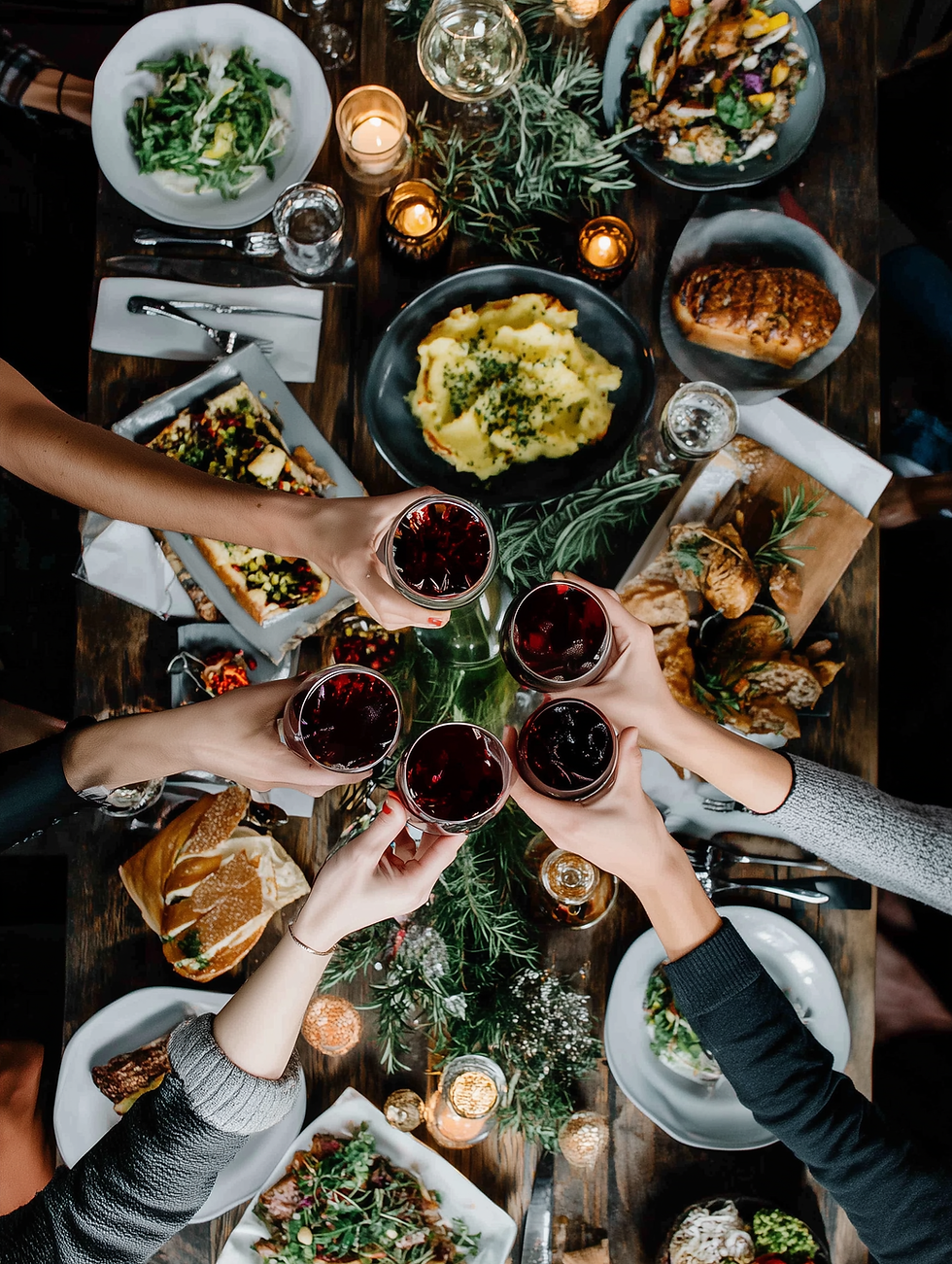 view looking down on a dining table full of food, candles, glasses, serving dishes and people reaching their hands in to cheers with glasses of wine