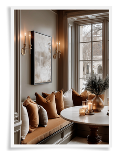 elegant dining niche with built in banquette seating, warm amber colored pillows, taupe walls, with art on the wall and sconces flanking the art. there is a winter scene outside the window and candles burning on top the dining table