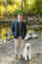 High school senior photo in a black jacket smiles beside a fluffy dog by a rocky stream. golden hour senior photo by a creek in Columbus, Ohio