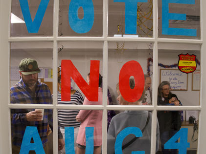 Community members gather inside a building displaying large “Vote No Aug 4” signage, promoting voter registration and hours of operation.