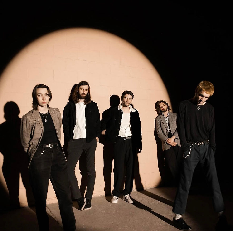 Social Cinema stands in a semi-circle under a dramatic spotlight against a backdrop of a peach-colored wall. Their casual yet stylish attire creates a moody and artistic atmosphere, with shadows adding depth to the scene.