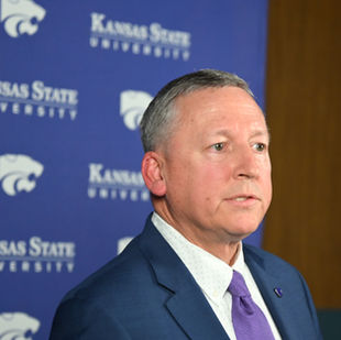 President Linton speaks to the press after his State of the University address, dressed in a formal suit and tie, with the university's logo prominently displayed on the backdrop.