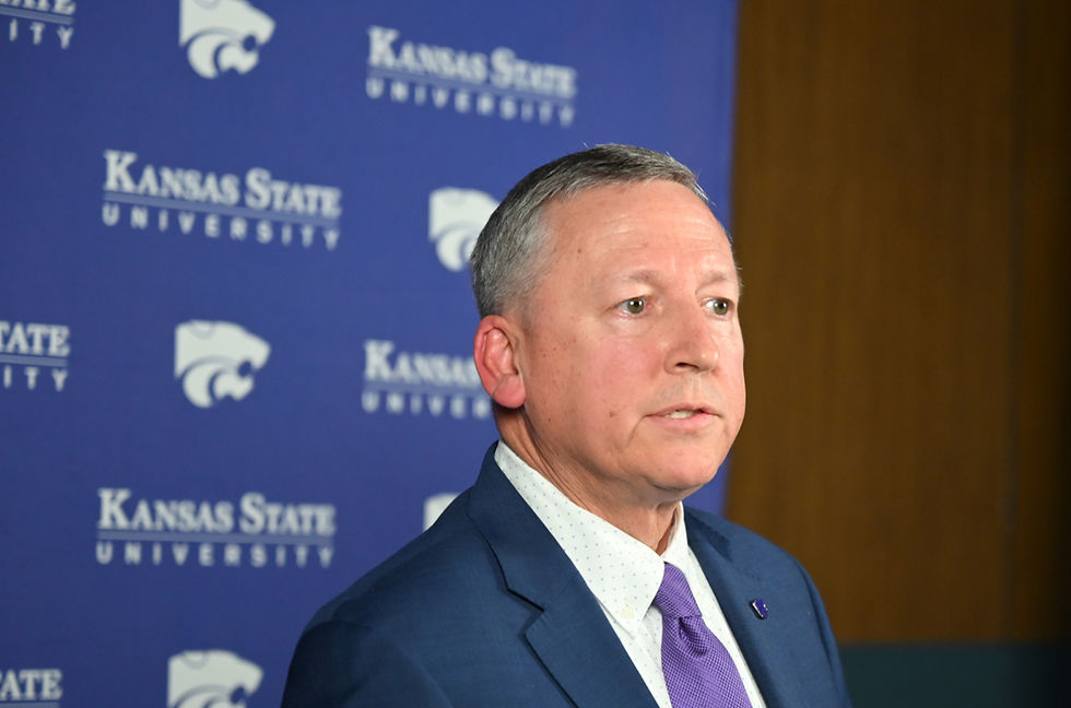 President Linton speaks to the press after his State of the University address, dressed in a formal suit and tie, with the university's logo prominently displayed on the backdrop.
