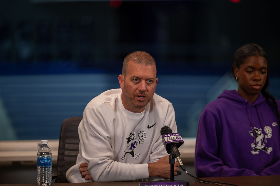Coach Mansfield speaks at a press conference, addressing a microphone, with an athlete seated next to him, both wearing K-State apparel.