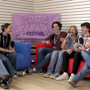 Woodridge sits on a red couch next to Cadence and Delaney on a blue couch with a Frog Wizard Festival banner in the middle. 