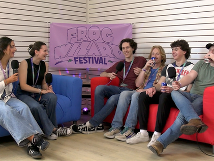Woodridge sits on a red couch next to Cadence and Delaney on a blue couch with a Frog Wizard Festival banner in the middle. 