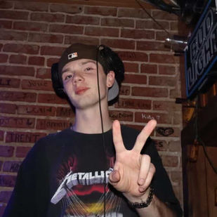 Jake Wright poses in a dimly lit bar, flashing a peace sign while wearing a Metallica shirt and a backward cap. 