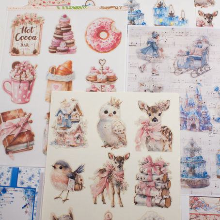 A flat lay photograph of various printable winter-themed junk journal stickers and fussy cuts. The images include a hot cocoa bar, ice castles, polar bears, and woodland animals. These are shown printed on a variety of surfaces including white cardstock, translucent vellum paper, a manila file folder, and a piece of vintage faded sheet music.