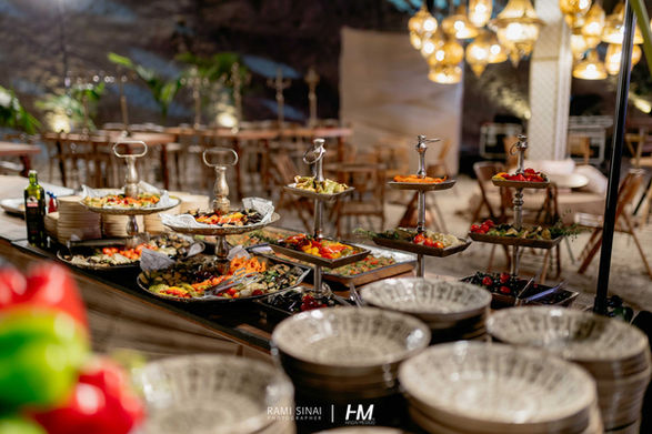 תקריב על מנורת שולחן דקורטיבית עם אהיל בד ופרנזים בגוון נחושת חם. ברקע, קירות הסלע המלאכותיים מוארים בהקרנות וידאו-ארט גיאומטריות בצבע תכלת, המייצרות ניגוד ויזואלי בין התאורה הקלאסית לאפקטים המודרניים. הפקה: חגי מגידו – HM Productions.