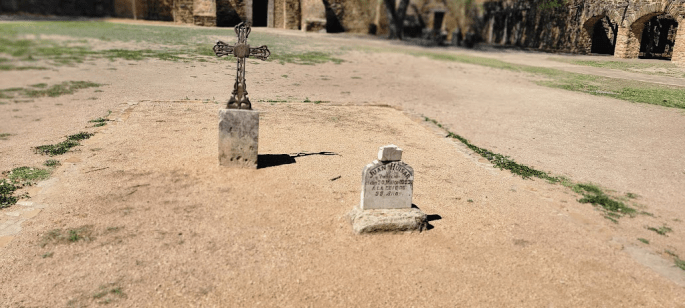 A grave site featuring a metal cross and a tombstone on a sandy ground, surrounded by grassy areas and ruins in the background.