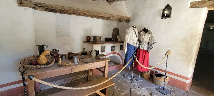 A historic kitchen setting featuring a wooden table with pottery, utensils, and a woven basket. Two mannequins are dressed in traditional clothing, with a red skirt and patterned shawl. The room has whitewashed walls, exposed wooden beams, and stone flooring.