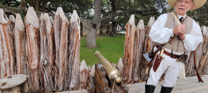 A historical reenactor dressed in period clothing stands beside a wooden fence and a cannon in a grassy area, surrounded by trees.