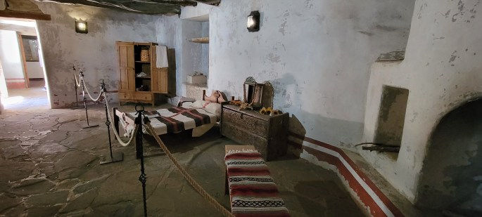 Interior of a rustic room featuring a simple bed with striped bedding, a wooden cabinet, and an intricately carved chest. The room has stone flooring, light-colored walls with some wear, and is illuminated by a wall light.