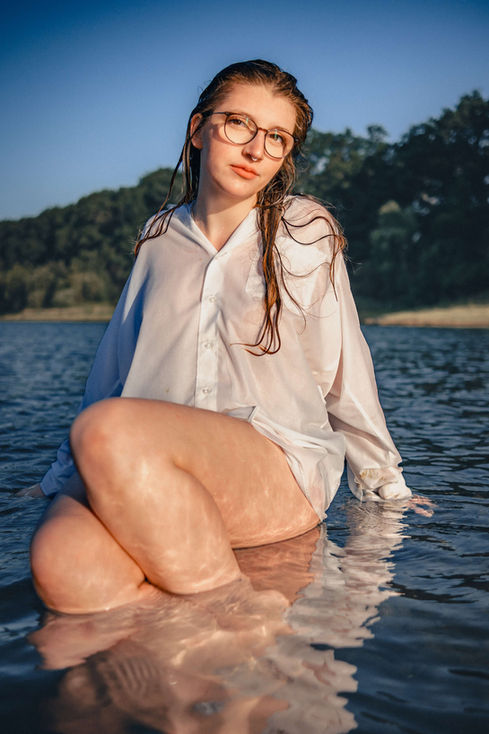 Junge Frau sitzt im Wasser, trägt Brille und weißes Hemd. Portrait Shooting.