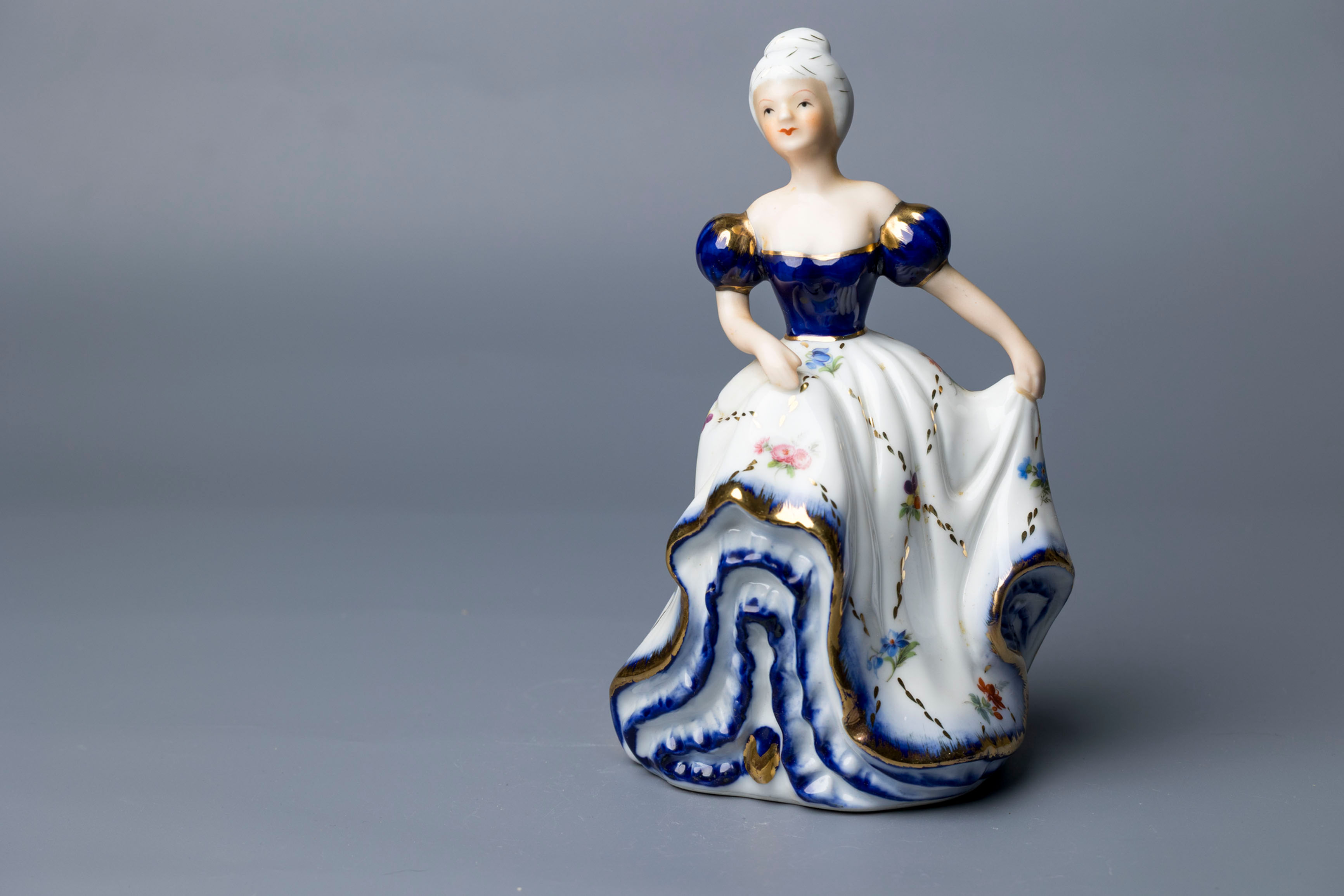 This is a hand-painted porcelain figurine of a woman in a full gown, featuring cobalt blue and gilded accents with floral