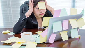 Stressed woman in an office with post-it notes on her face and laptop. Wooden desk, coffee cup nearby. Bright, busy environment.