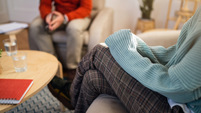 Two people sitting in a cozy room with a wooden table. One wears a light blue sweater, plaid pants; the other in red. Notebook, glasses on table.