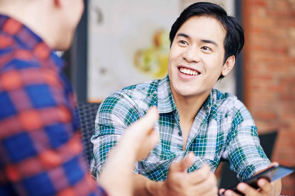 Smiling man in a green plaid shirt holds a phone, talking to another person in a blue plaid shirt. Bright, casual setting with brick wall.